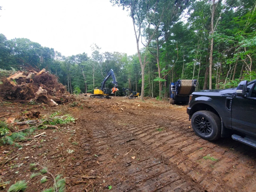 Excavators clearing a wooded lot, with a pile of stumps and brush beside a grinder at the tree line.