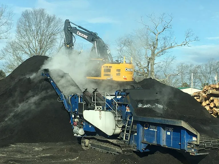 Excavator loading a grinder beside steaming piles of compost at the yard, with stacked logs behind.